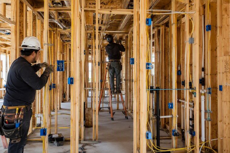Electricians running cable to boxes through open stud bays, with the plumbing and heating already in